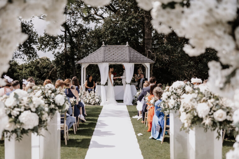 Outdoor wedding ceremony at Trunkwell House in Berkshire with the couple exchanging vows beneath a white gazebo, surrounded by guests and white floral arrangements.